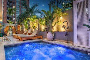 A rooftop pool area with lounge chairs, a hot tub, and tropical plants.