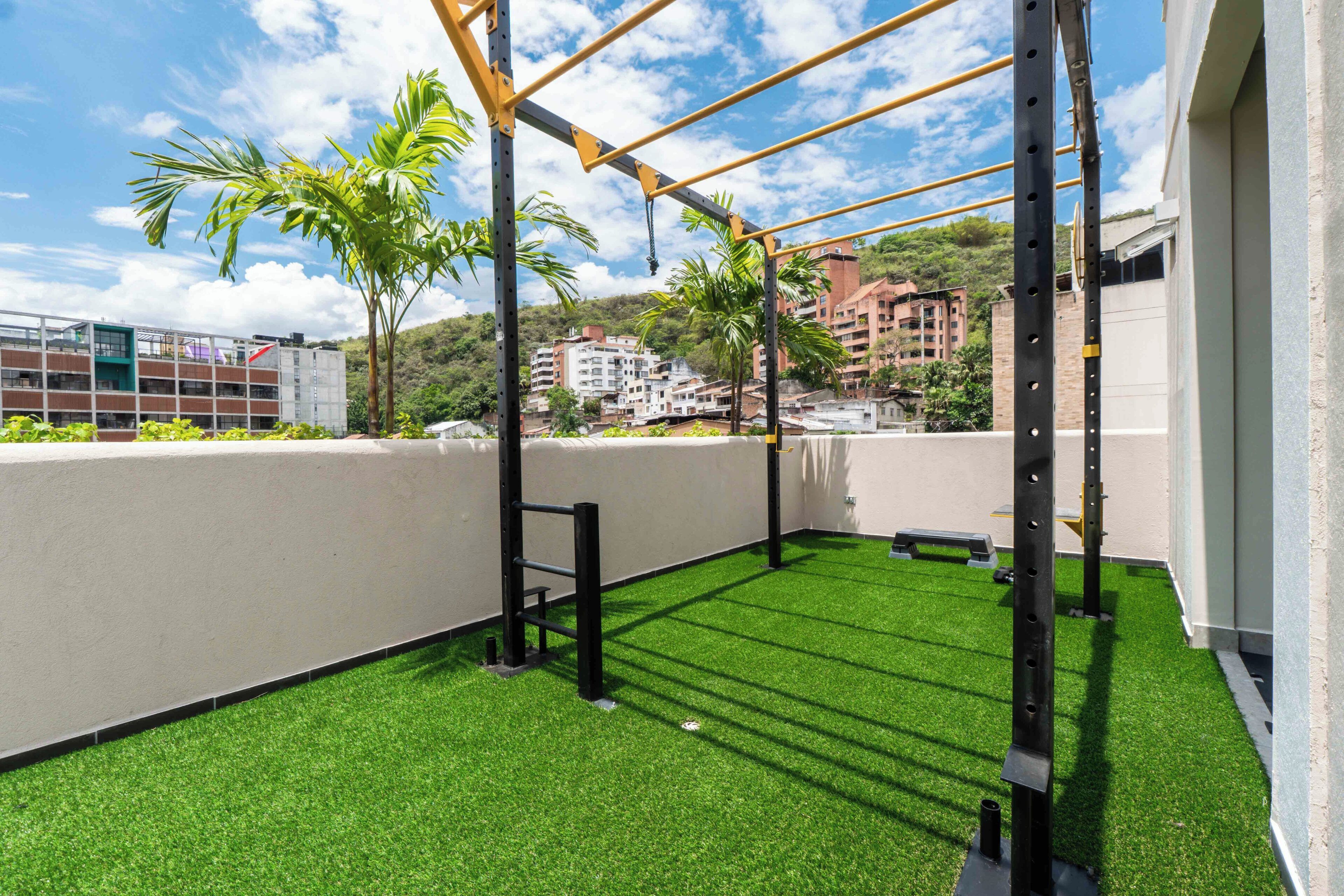 Espaço de ginástica ao ar livre com grama sintética, equipamentos de exercício e vista para edifícios próximos.