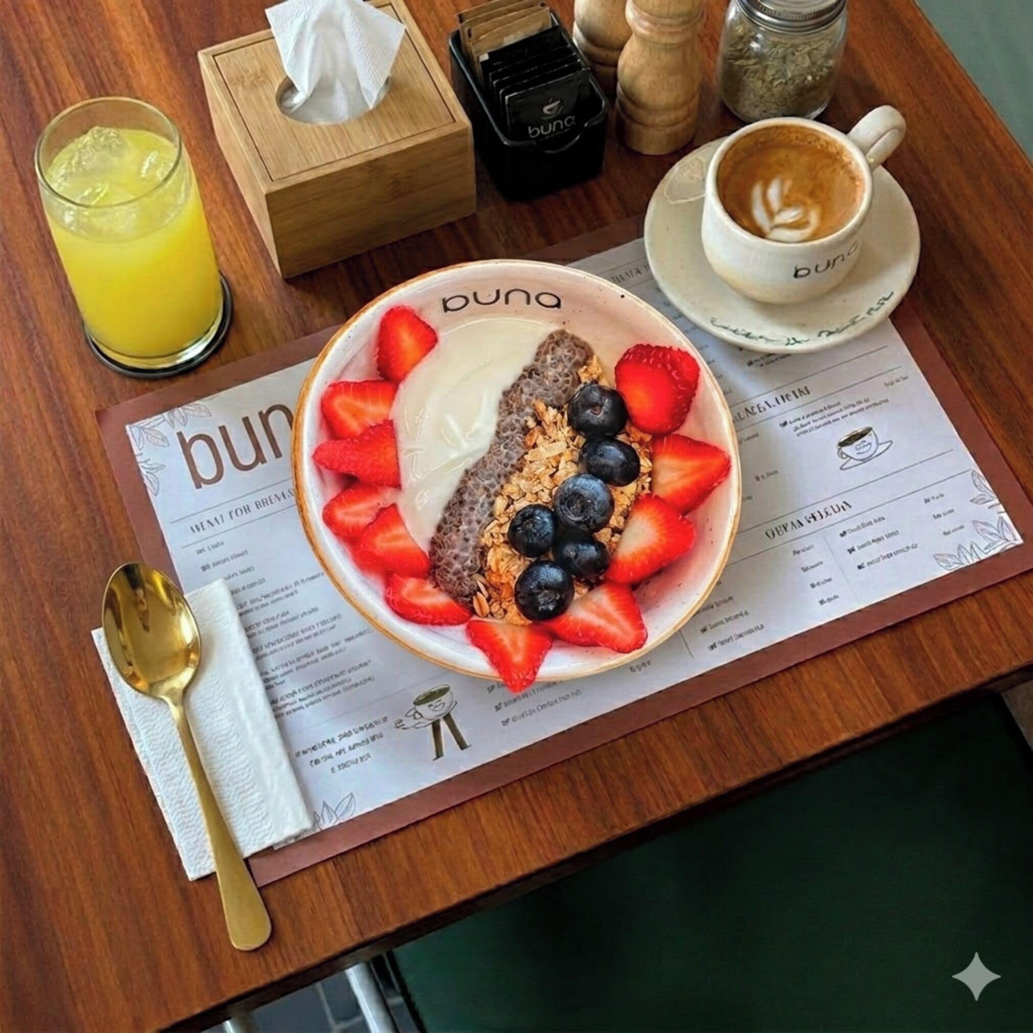 Un desayuno con un plato de yogur, fresas, arándanos y granola, un vaso de zumo de naranja, una taza de café, una cuchara, una servilleta, una caja de pañuelos y un menú.