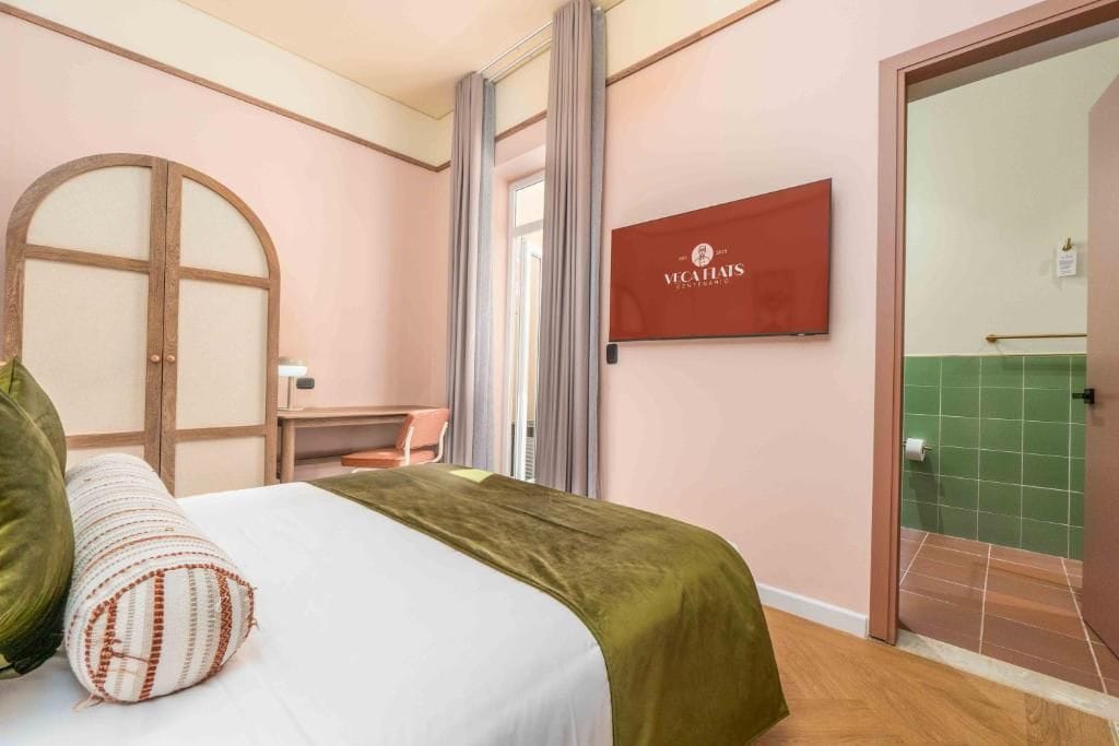 A hotel room with a bed, a desk, a chair, a large mirror, and a door leading to a bathroom.