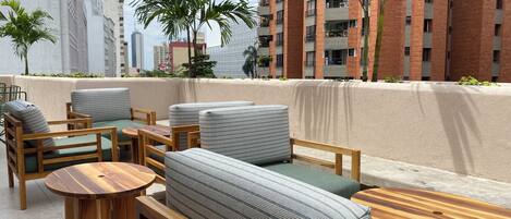 Terraza en la azotea con mobiliario exterior, incluyendo sofás y mesas, rodeada de edificios altos y algo de vegetación.
