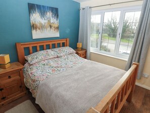 Een slaapkamer met een houten bed, bloemenbedrukking, een nachtkastje met een lamp, een dressoir en een raam met uitzicht.