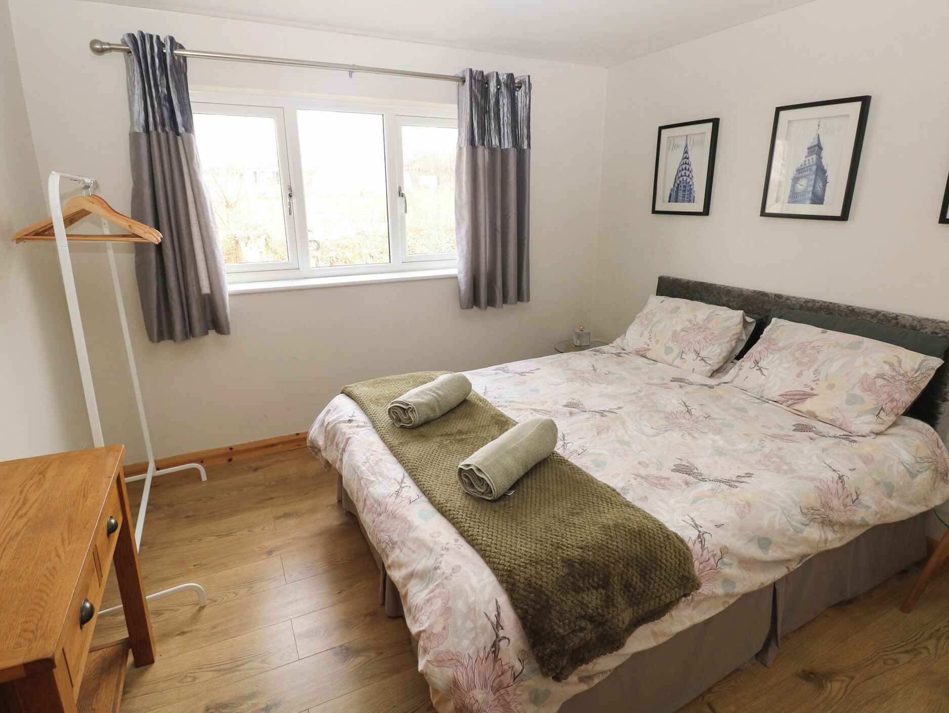 Une chambre à coucher avec un lit, un bureau en bois, une fenêtre avec des rideaux et deux tableaux encadrés accrochés au mur.