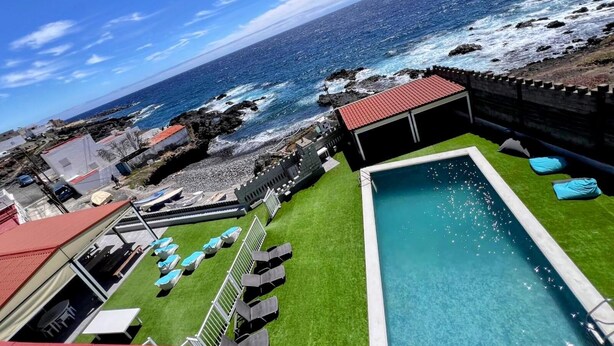 Une piscine avec vue sur l’océan, un bâtiment à toit rouge et une pelouse verdoyante agrémentée de chaises longues.