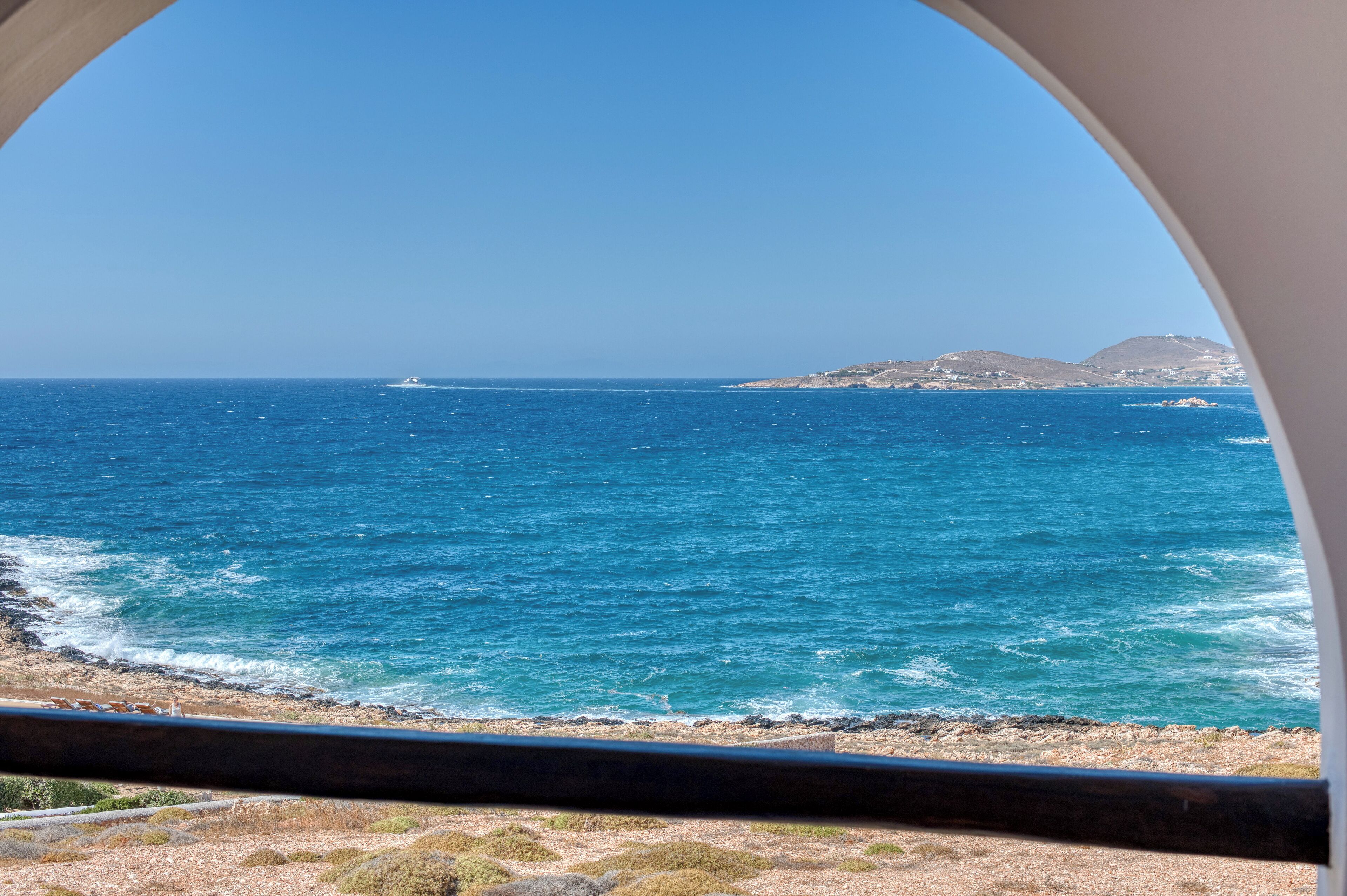 A coastal view through a circular window, featuring the sea, a distant boat, and a rocky shoreline with sparse vegetation.