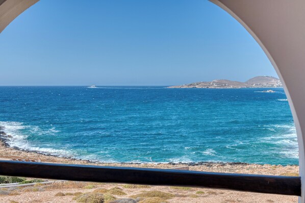 A coastal view through a circular window, featuring the sea, a distant boat, and a rocky shoreline with sparse vegetation.
