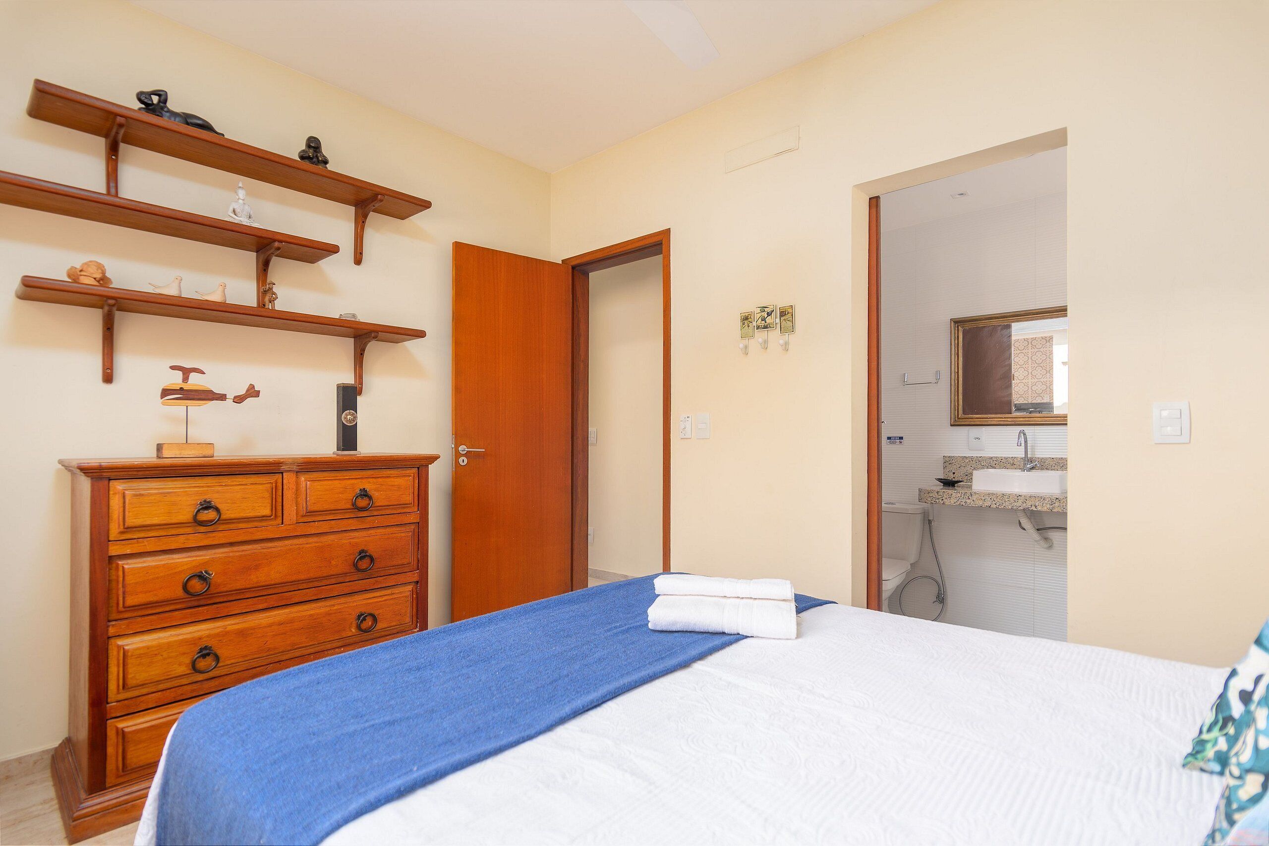 Quarto com cama, cómoda de madeira e banheiro visível por uma porta aberta.