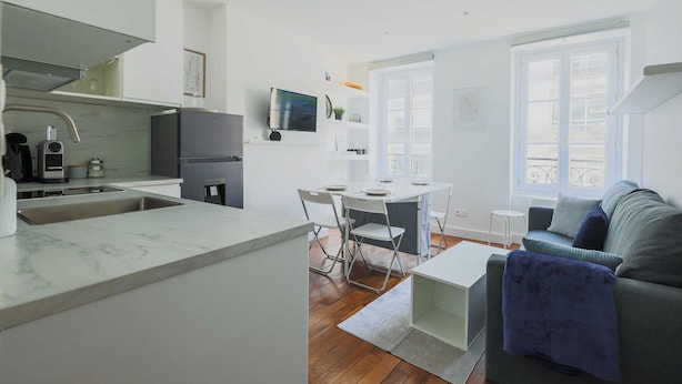 Een moderne keuken met een marmeren aanrecht, een eethoek met een witte tafel en stoelen, en een grijze bank in een lichte woonkamer met grote ramen.