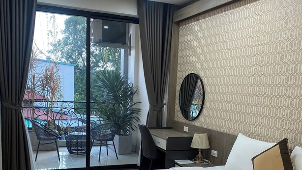 A hotel room with a large bed, a desk with a chair, a small table with a lamp, and a balcony with plants. - Royal Lotus Apartment and Hotel Ha Noi (Hanoi)