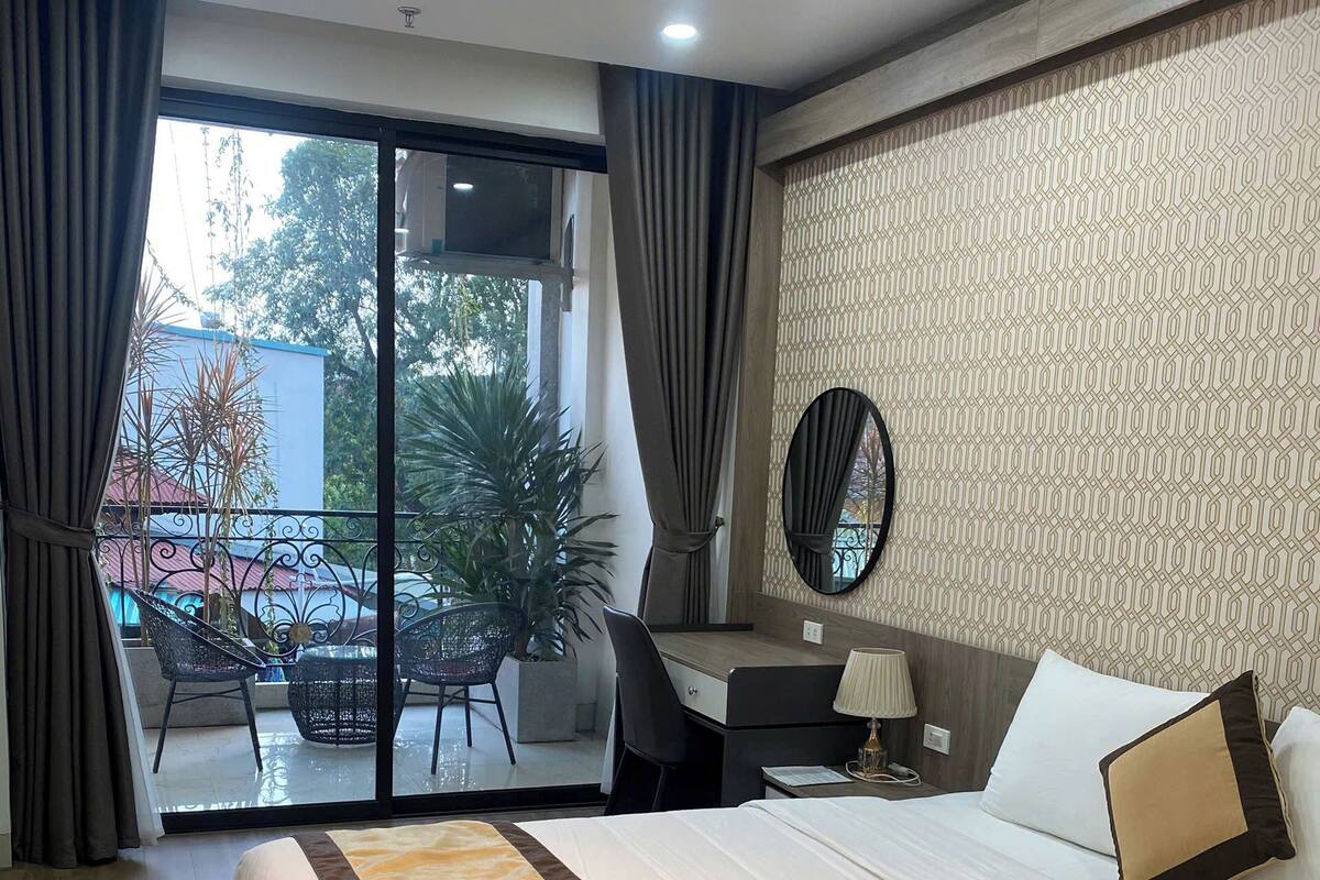A hotel room with a large bed, a desk with a chair, a small table with a lamp, and a balcony with plants.