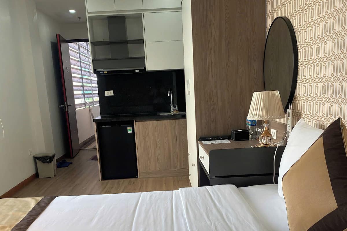 A modern hotel room with a large bed, a small kitchenette, and a bedside table with a lamp.