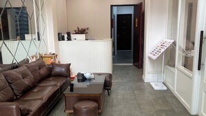 A modern living room with a brown leather sofa, a coffee table, and a small ottoman. There is a reception desk with a sign and a display stand with information. - Royal Lotus Apartment and Hotel Ha Noi (Hanoi)