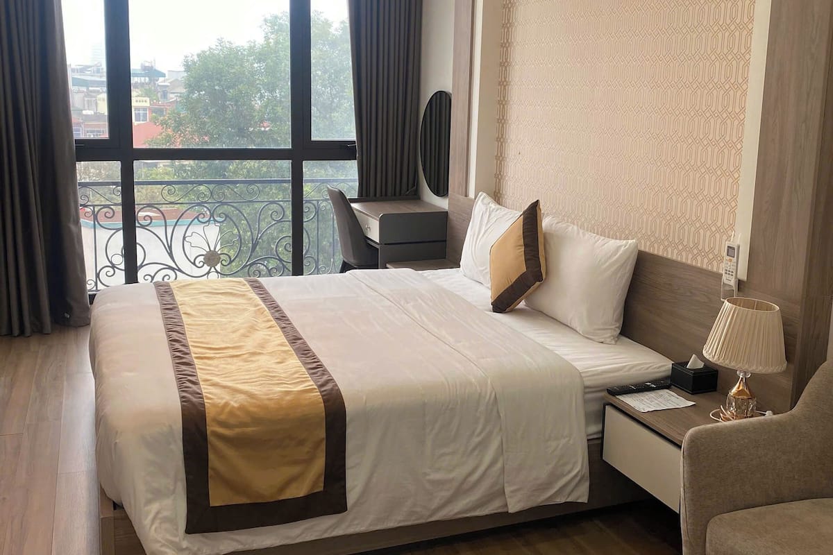 A hotel room with a large bed, a bedside table with a lamp, a chair, and a view of the city through large windows.