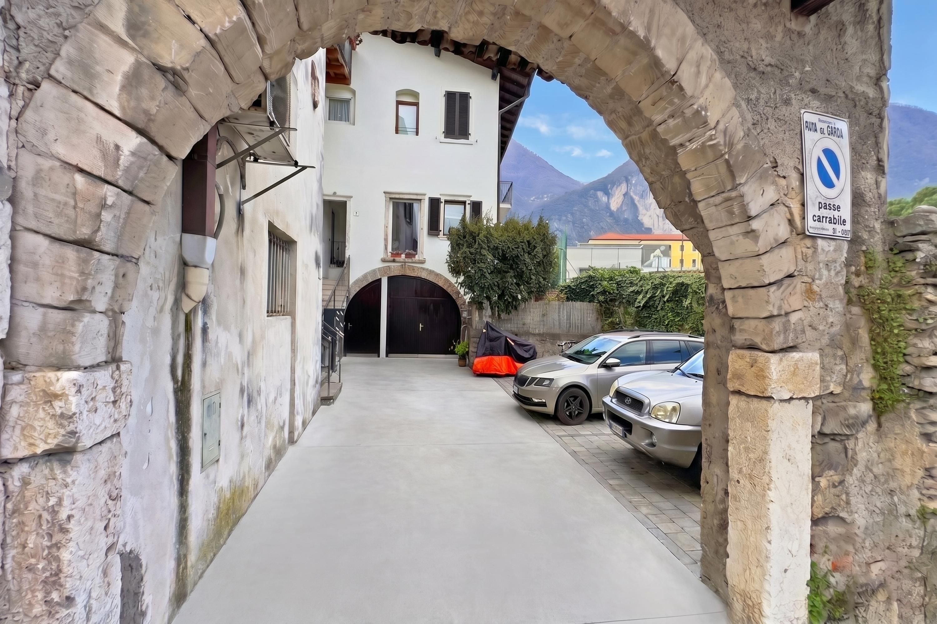 A narrow street with a stone archway, a building with a black gate, and a sign indicating a no parking zone.