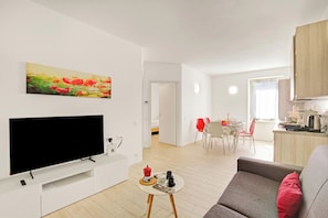 Een moderne woonkamer met een flatscreen televisie, een schilderij, een klein tafeltje met een rode kaars, en een grijze bank.