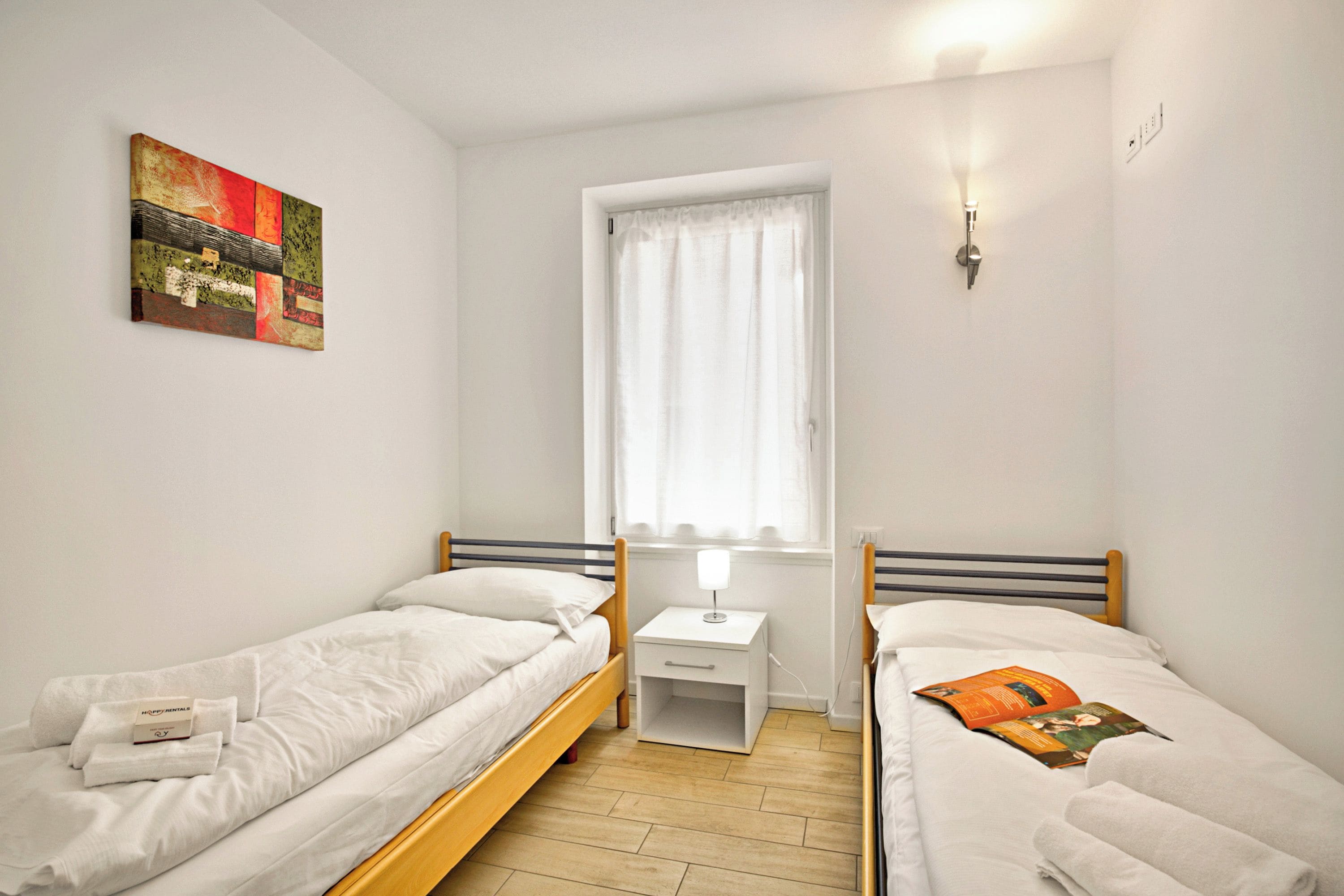 Ein Zimmer mit zwei Einzelbetten, einem Nachttisch mit Lampe, einem Fenster mit Vorhängen und einem Bild an der Wand.