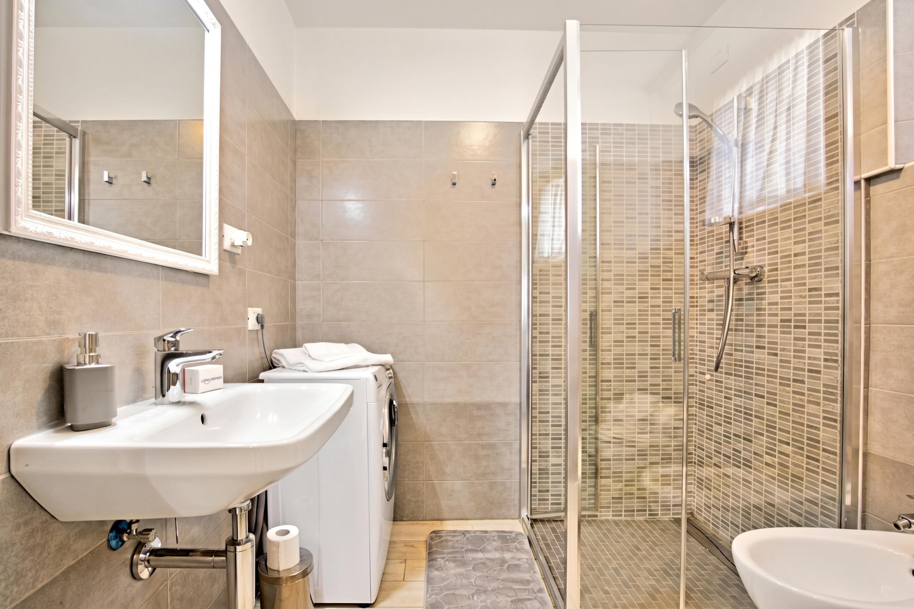 A modern bathroom with a large sink, a washing machine, a shower with glass doors, and a mirror.