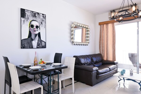 A dining area with a table set for two, a brown leather sofa, and a decorative mirror. - Camenzuli Apartments (Sliema)