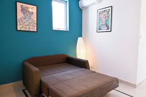 A room with teal walls, a brown sofa, two framed artworks, a window, and an air conditioning unit. - Camenzuli Apartments (Sliema)