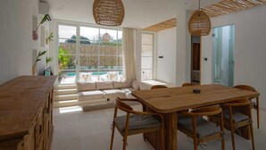 A spacious dining area with a wooden table and chairs, a sofa with cushions, and a view of a swimming pool through large windows.