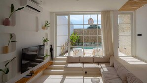 A modern living room with a large sectional sofa, a flat-screen TV, and a wooden coffee table. There are multiple potted plants and a view of a pool and outdoor area through large windows.