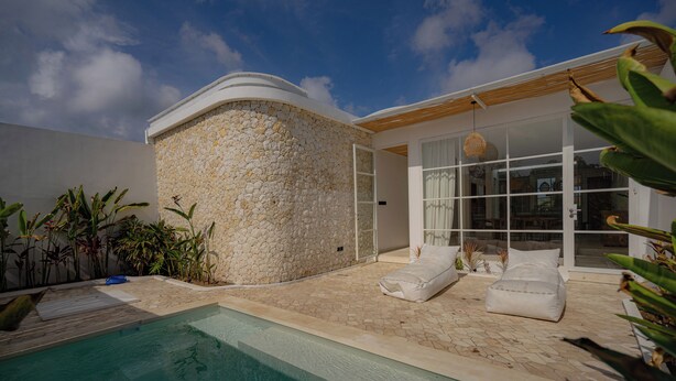 A poolside area with a stone wall, white lounge chairs, and a glass door leading to a covered patio.