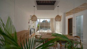A modern dining area with a wooden table and chairs, a woven pendant light, and a view of the outdoors through large windows.