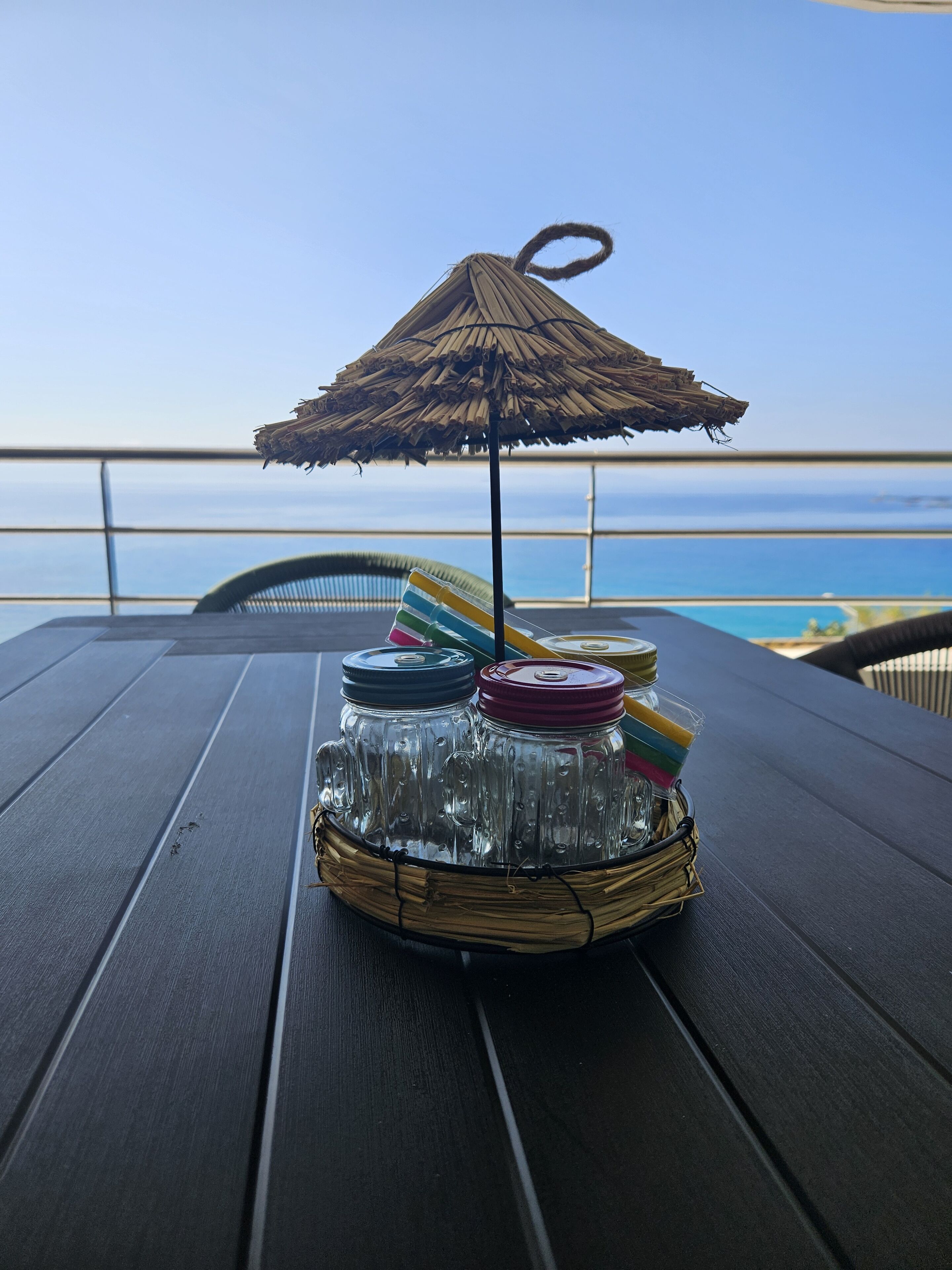 A table with a woven basket containing jars and a straw umbrella, overlooking a beach.