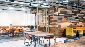 A modern dining area with wooden tables and chairs, a glass partition, and a wall with wooden blocks. - Chapter Lewisham Student Only Cozy Stay (London)