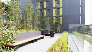 An outdoor seating area with benches and planters, surrounded by a modern building with large windows and a glass railing. - Chapter Lewisham Student Only Cozy Stay (London)