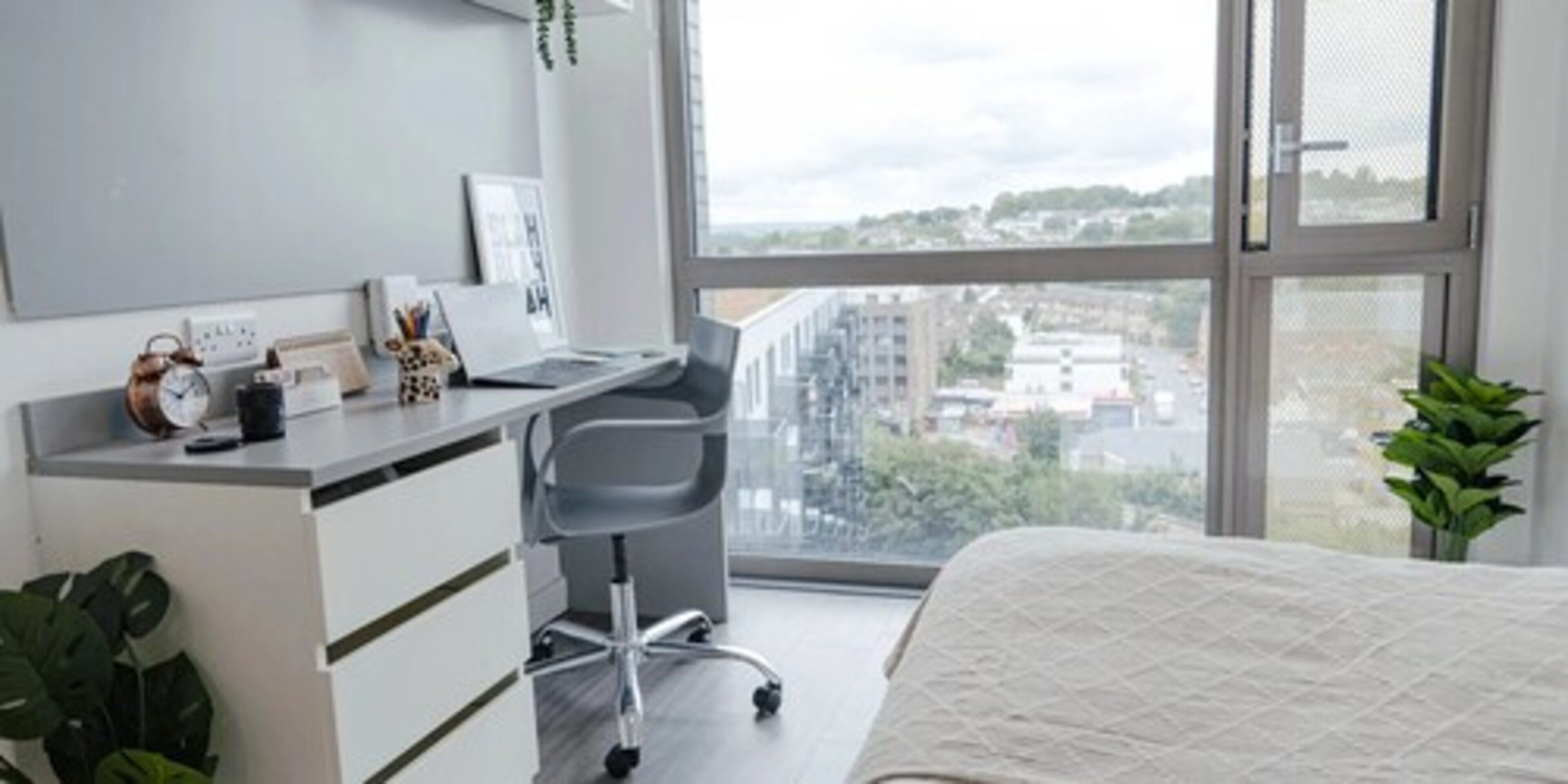 A modern bedroom with a bed, a desk with a chair, a window with a view of buildings, and a potted plant.