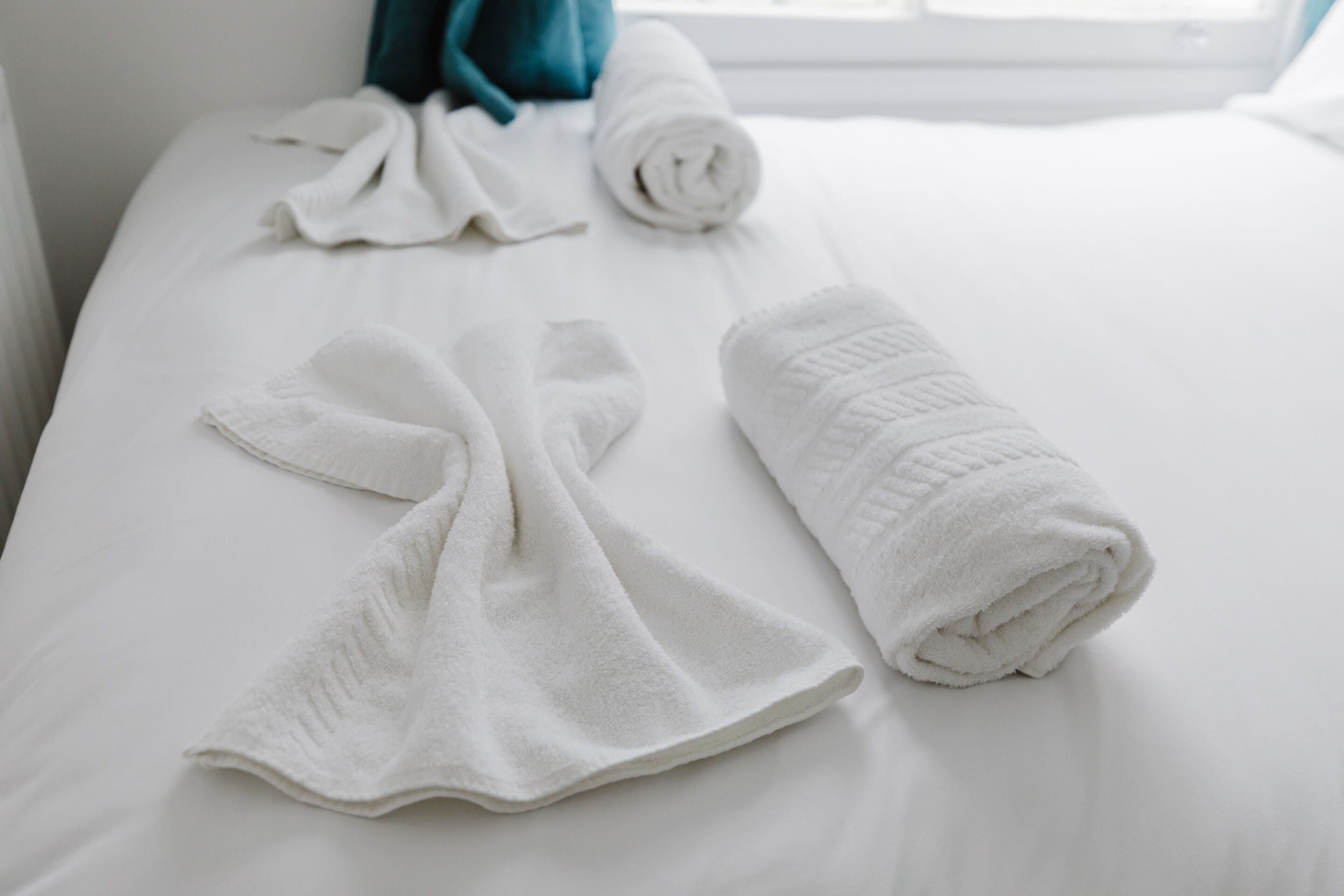 A neatly made bed with white linens and towels, a teal-colored pillow, and a window in the background.
