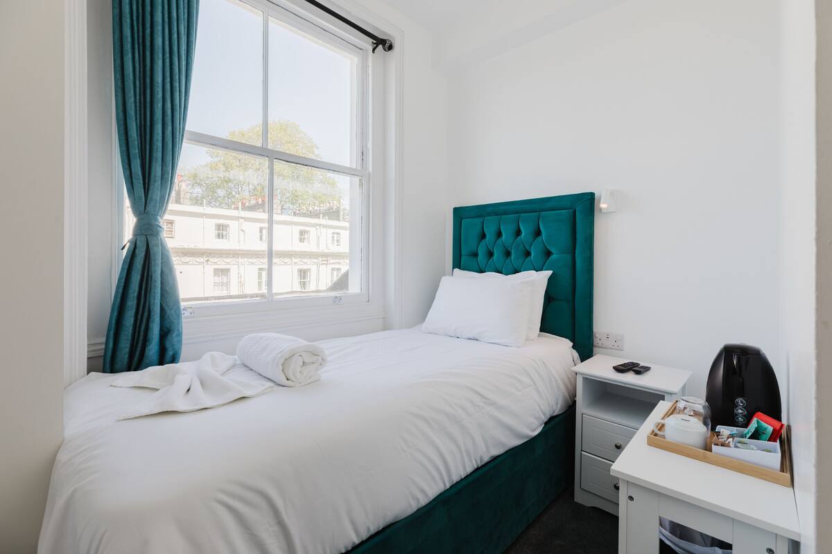 A neatly made bed with white linens, a teal headboard, a bedside table with a phone and a coffee maker, and a window with teal curtains.