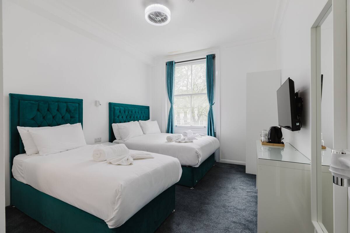 A hotel room with two beds, a TV, a mirror, and a window with curtains.