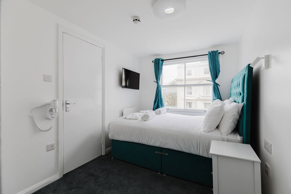 A bedroom with a bed, a TV mounted on the wall, a window with curtains, a door, and a power outlet.