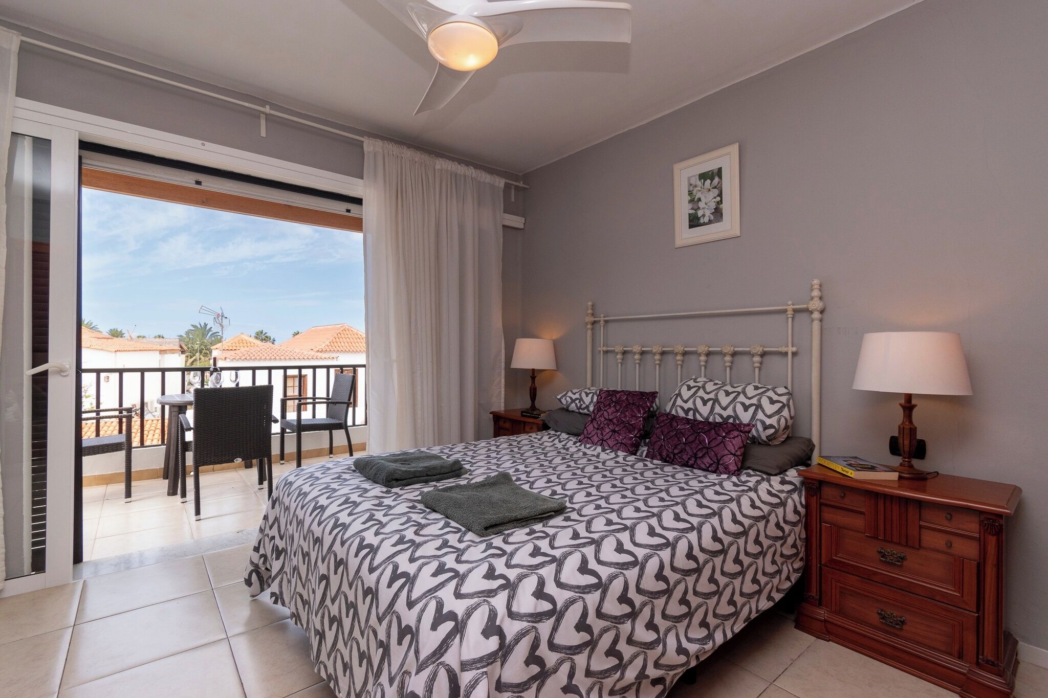 Un dormitorio con una cama grande, dos mesitas de noche con lámparas, un ventilador de techo y vistas al balcón exterior.