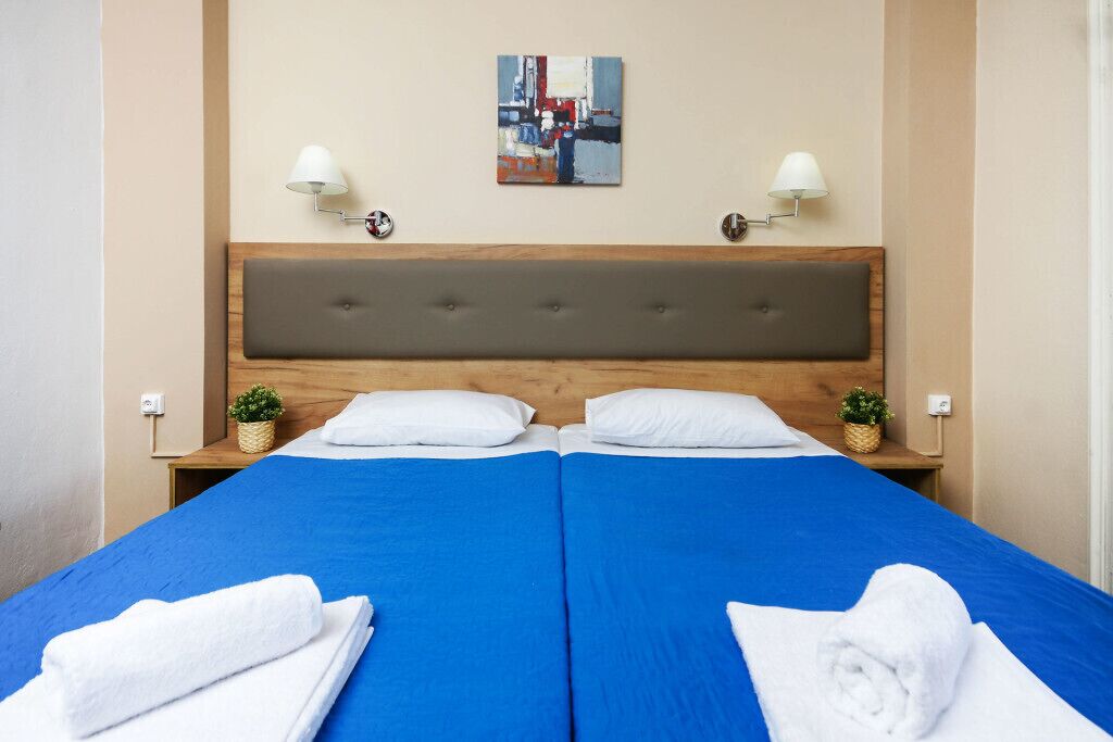 Una cama doble con ropa de cama azul, dos lámparas de noche, un cabecero y un cuadro en la pared.