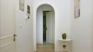 A narrow hallway with white walls, a tiled floor, a door on the left, a small table with a plant on the right, and a ceiling light. - Stavros Apartments Gouvia Marina (Corfu)