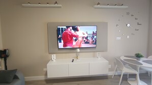 Una sala de estar moderna con un televisor de pantalla plana montado en la pared, un mueble blanco de entretenimiento debajo, un sofá gris y una pequeña mesa redonda con una silla.