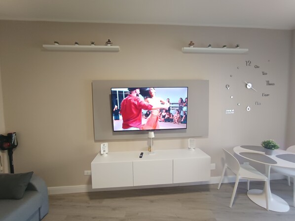 Una sala de estar moderna con un televisor de pantalla plana montado en la pared, un mueble blanco de entretenimiento debajo, un sofá gris y una pequeña mesa redonda con una silla.