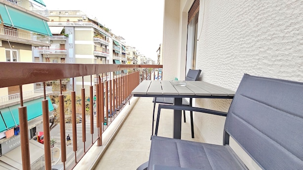 Un balcón con mesa y sillas, con vista a una calle con edificios y coches aparcados.