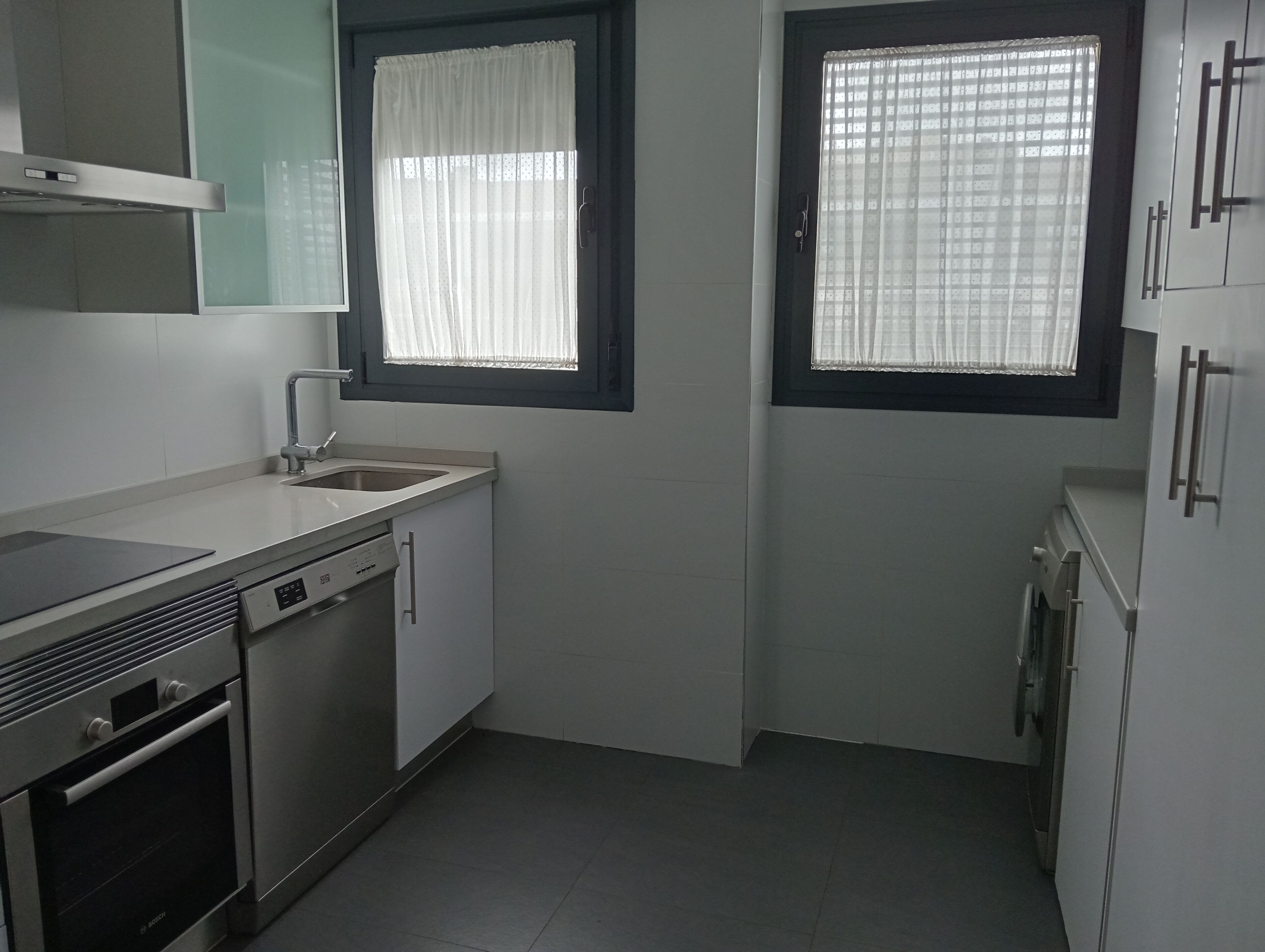 Una cocina moderna con horno empotrado, lavavajillas y un lavabo con grifo. Hay dos ventanas con cortinas, una sobre el lavabo y otra sobre la hornilla.