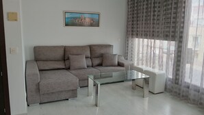 Un salon moderne avec un canapé gris, une table basse en verre et un fauteuil blanc. Un tableau représentant un paysage urbain, encadré, est accroché au mur.