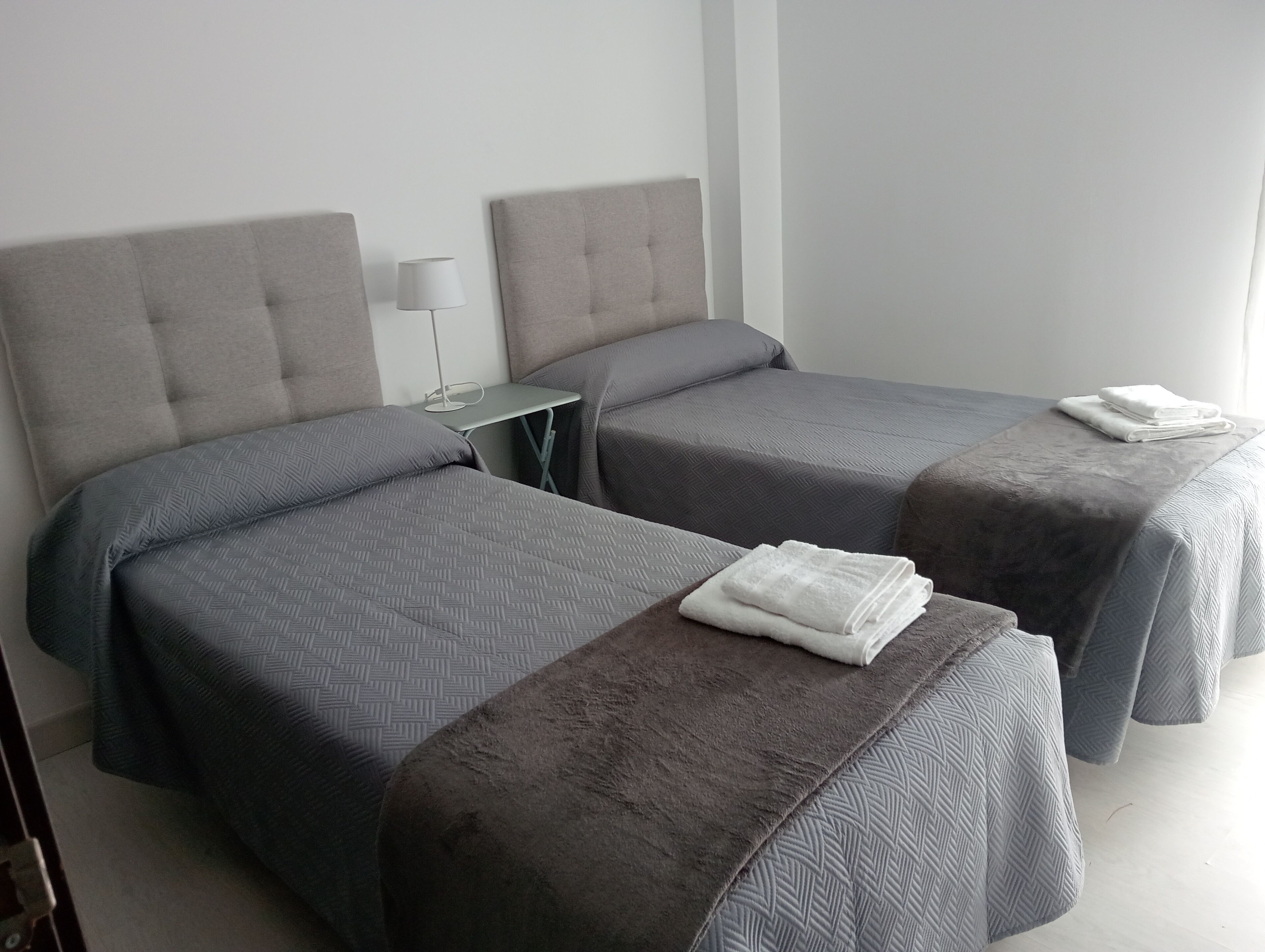 Habitación de hotel con dos camas, cada una con cabecero gris y ropa de cama a juego. Hay una mesita con lámpara entre las camas.