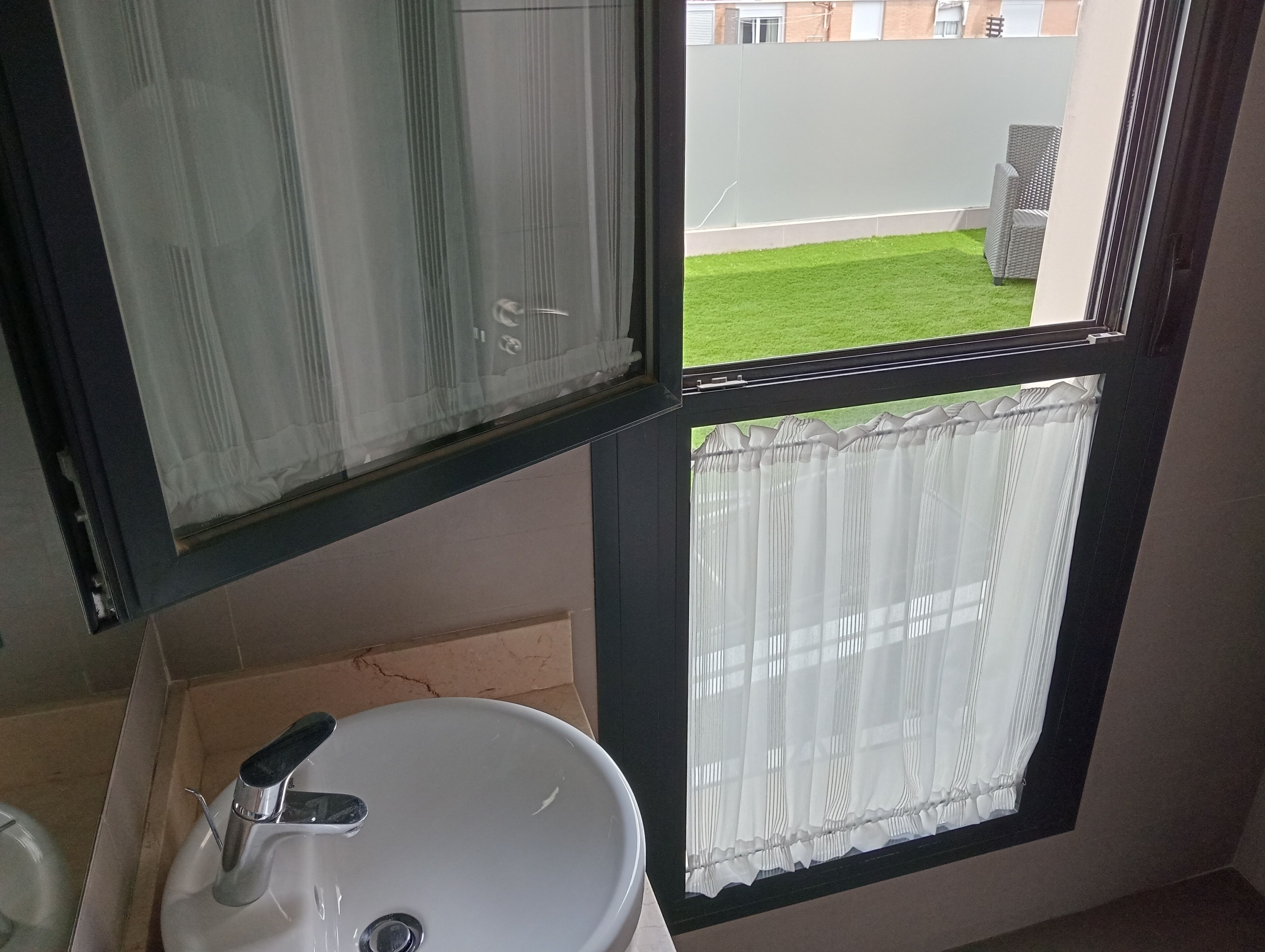 Un baño con un lavamanos blanco, un espejo y un ventanal con vistas a un césped verde y una pared blanca.