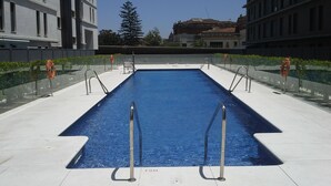 Une piscine rectangulaire avec des garde-corps en métal, entourée d’une terrasse carrelée, avec des bâtiments et un arbre en arrière-plan.