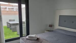 Une chambre à coucher comprenant un lit, une table de chevet, une lampe et donnant sur une terrasse extérieure aménagée avec du mobilier.