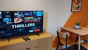 A modern living room with a flat-screen TV displaying a Netflix interface, a wooden cabinet with drawers, a dining table with chairs, and a potted plant.
