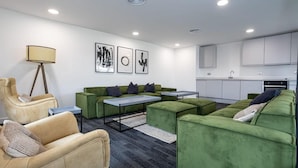 A modern living room with green sofas, a beige armchair, and a small coffee table. There is a kitchen area with white cabinets and a sink.