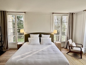 Une chambre d’hôtel avec un grand lit, deux tables de chevet, une chaise et de grandes fenêtres avec des rideaux.
